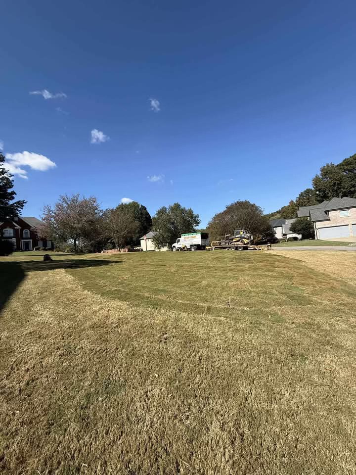 A wide grassy field with houses and trees under a blue sky.